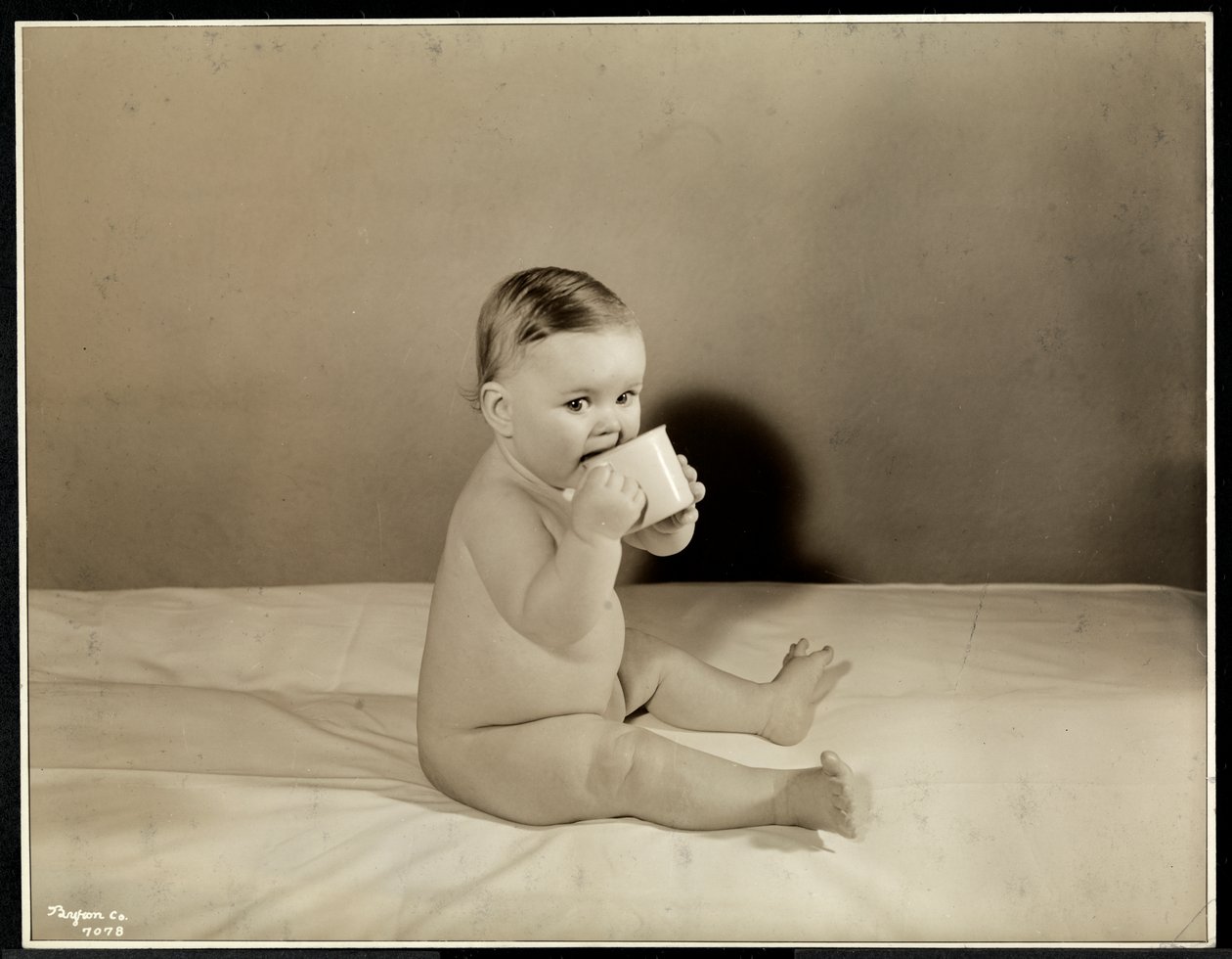 Ein Baby sitzt im Bett und trinkt aus einer Tasse im Maternity Center Association an der 70th Street East River, New York, 1935 von Byron Company