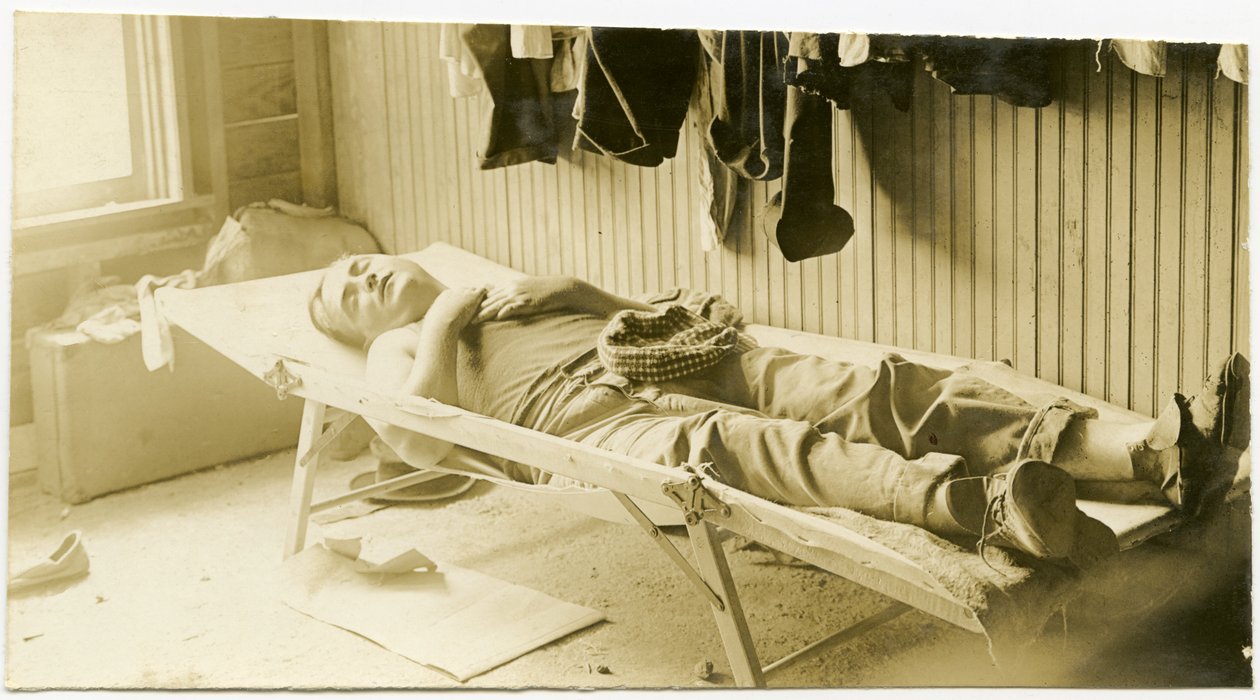Horizontal, Sepia-Fotografie zeigt einen Mann, der auf einem Feldbett in einem holzgetäfelten Gebäude schläft. Verschiedene Kleidungsstücke hängen an der Wand, und Koffer stehen in der Ecke des Raumes auf dem Boden von Carl Michel