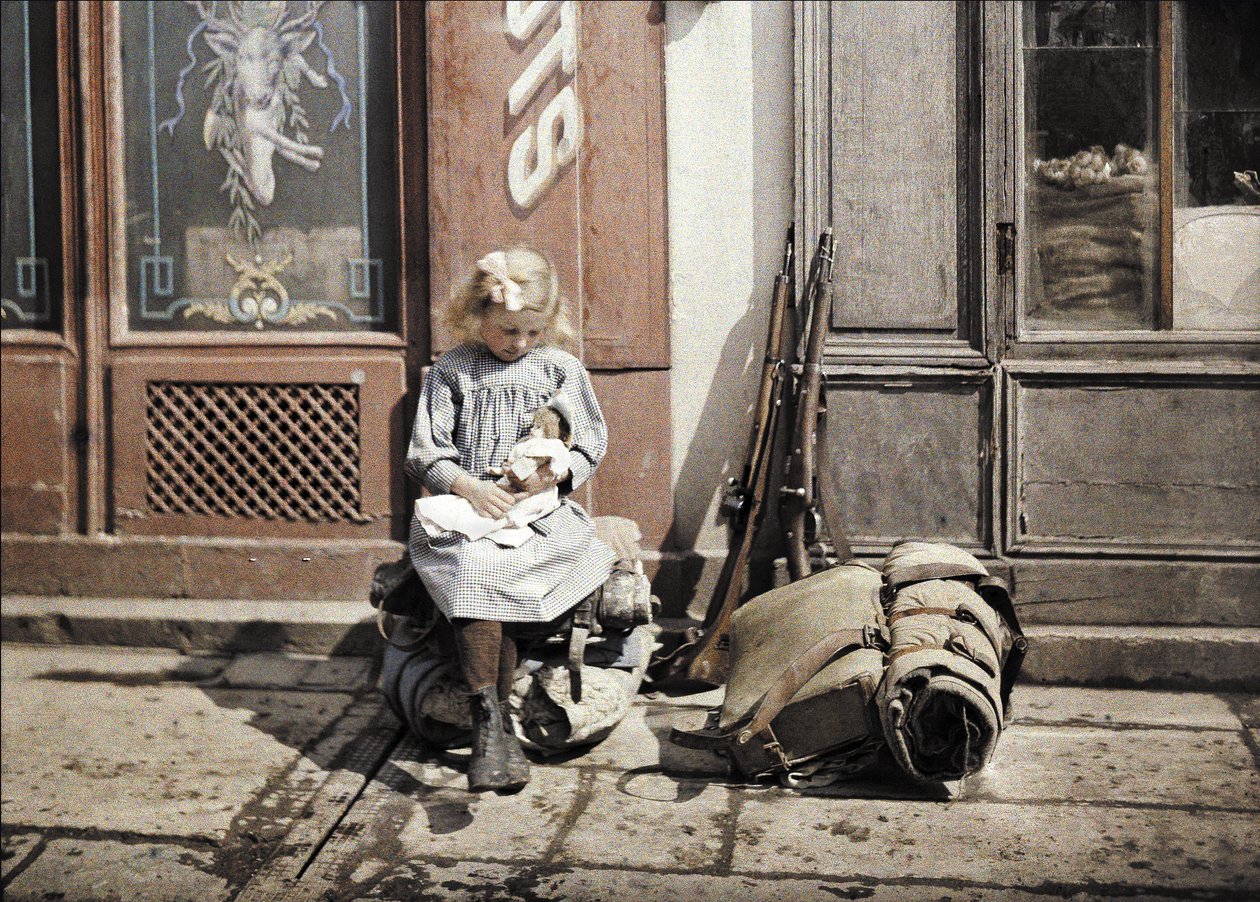 Ein kleines Mädchen spielt mit ihrer Puppe; zwei Gewehre und ein Rucksack liegen neben ihr auf dem Boden, Reims, Marne, Frankreich, 1917 (Autochrom) von Fernand Cuville